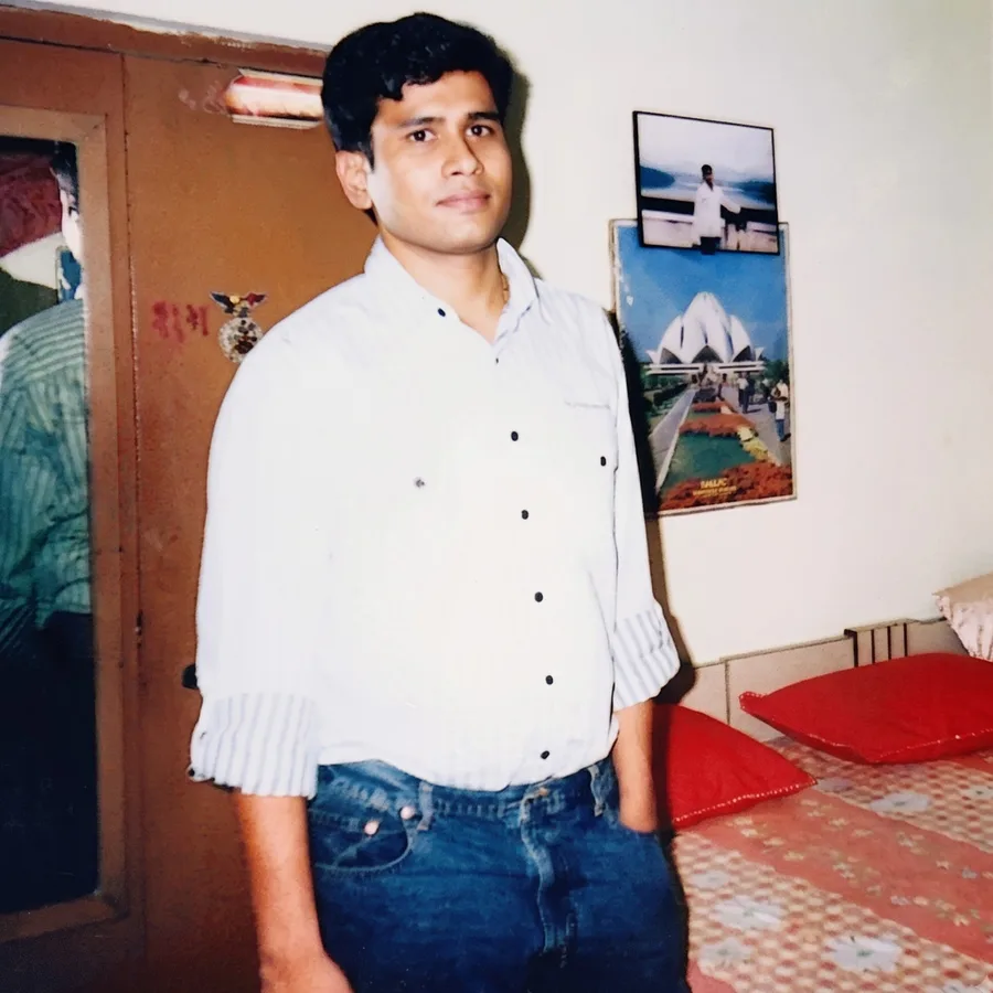An old print: a young Lavi in a white shirt and jeans, standing in a bedroom with framed pictures on the wall.