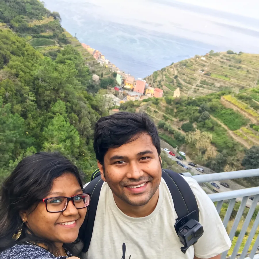 A selfie of Lavi and his wife on a cliffside path above a coastal village and terraced vineyards.