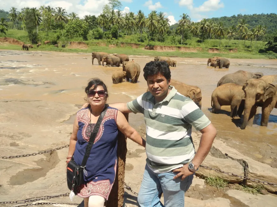 Lavi and a family member on a riverbank, with a herd of elephants bathing in the river behind them.