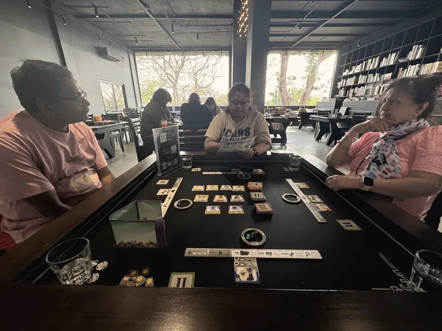 Family around a dark wooden game table in a cafe, tiles laid out mid-game, with shelves of board games behind them.