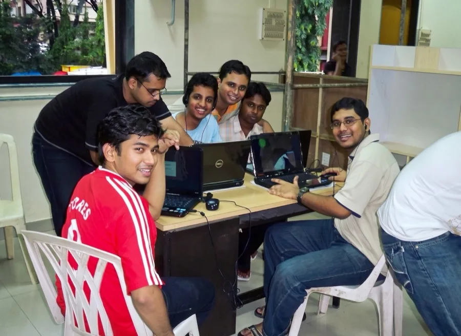 A group of classmates gather around laptops at a table, smiling back at the camera.