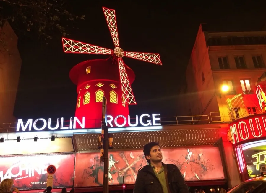 Lavi looks up in front of the glowing Moulin Rouge windmill at night.