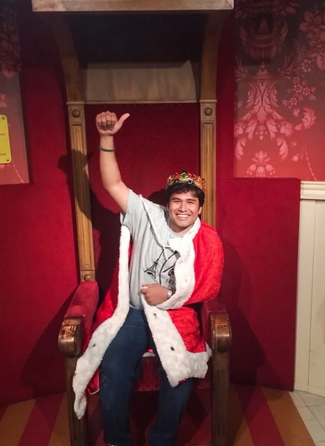 Lavi poses in a crown and robe on a red throne, one arm raised.