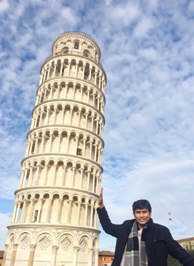 Lavi leans against the Leaning Tower of Pisa under a cloudy sky.