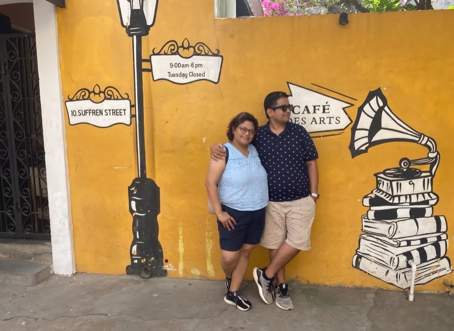 Lavi and a woman lean together against a yellow cafe wall with a gramophone mural.