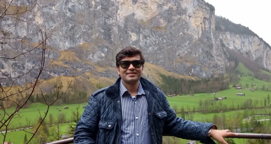 Lavi stands at a railing in front of a green valley and rocky cliff.