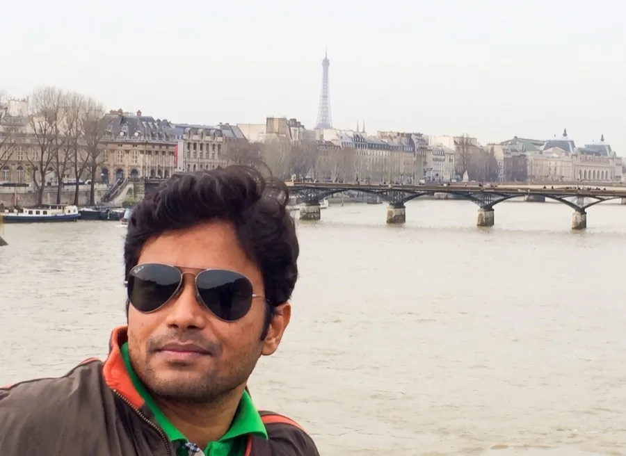 Lavi takes a selfie in sunglasses by a river with the Eiffel Tower in the distance.