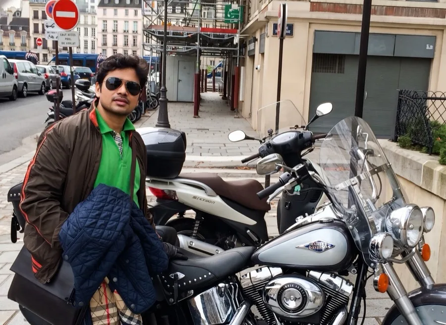 Lavi poses in sunglasses beside a parked motorcycle on a Paris street.