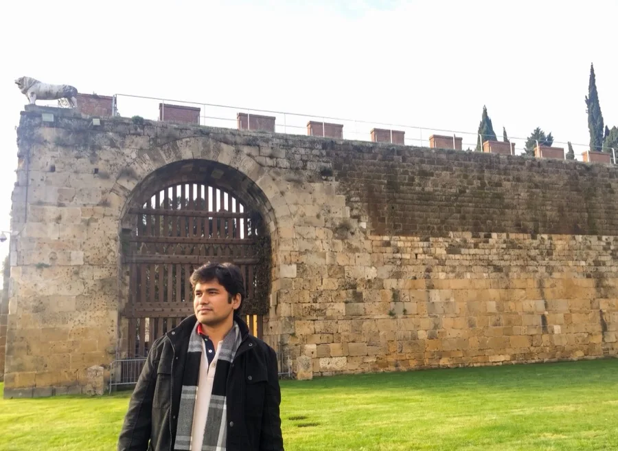 Lavi stands in front of a stone city wall with an arched gate.