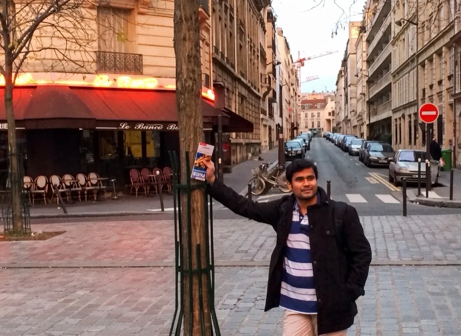 Lavi leans on a tree on a quiet Paris street lined with cafes.