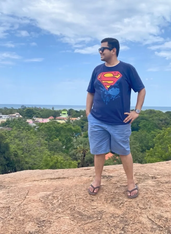 Lavi stands on a rocky hilltop in a Superman tee above green trees.