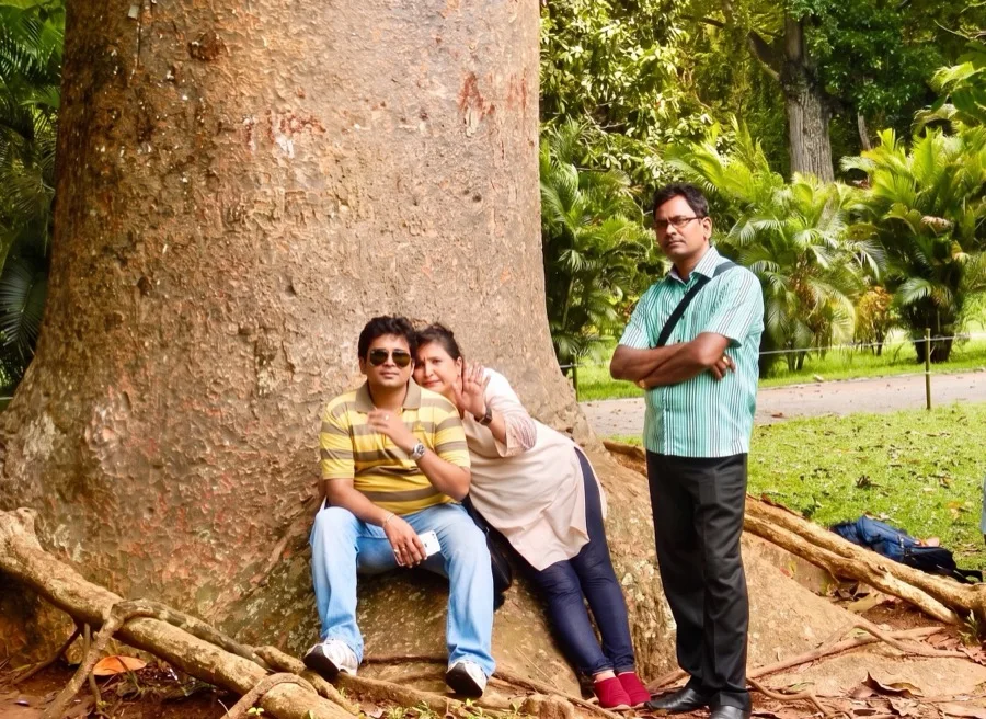 Lavi stands beside two people sitting at the roots of a huge tree.