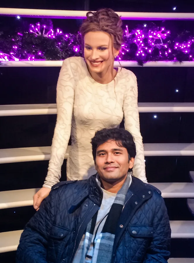 Lavi sits in a quilted jacket in front of a smiling wax figure under purple lights.