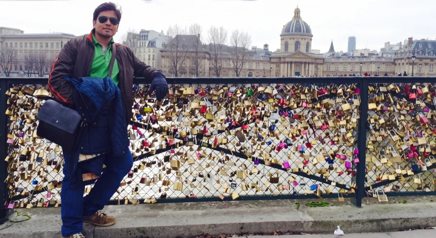 Lavi in sunglasses and a brown jacket leans on a railing covered in love locks beside the Seine, with a domed Paris building behind.