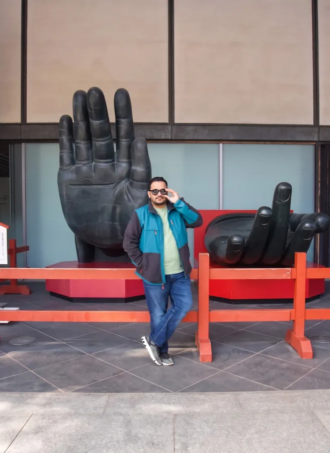 Lavi in sunglasses and a teal jacket stands between two giant black hand sculptures on a red platform.