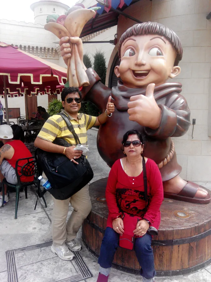 A man in a yellow striped shirt and a woman in red pose with a large cartoon monk statue.