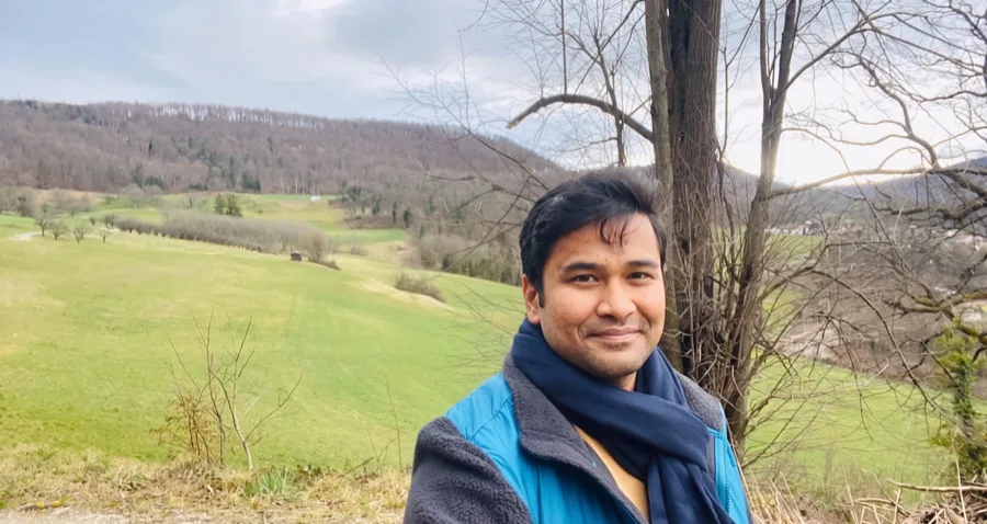 Lavi in a blue jacket and scarf smiles in front of a green valley and bare trees.